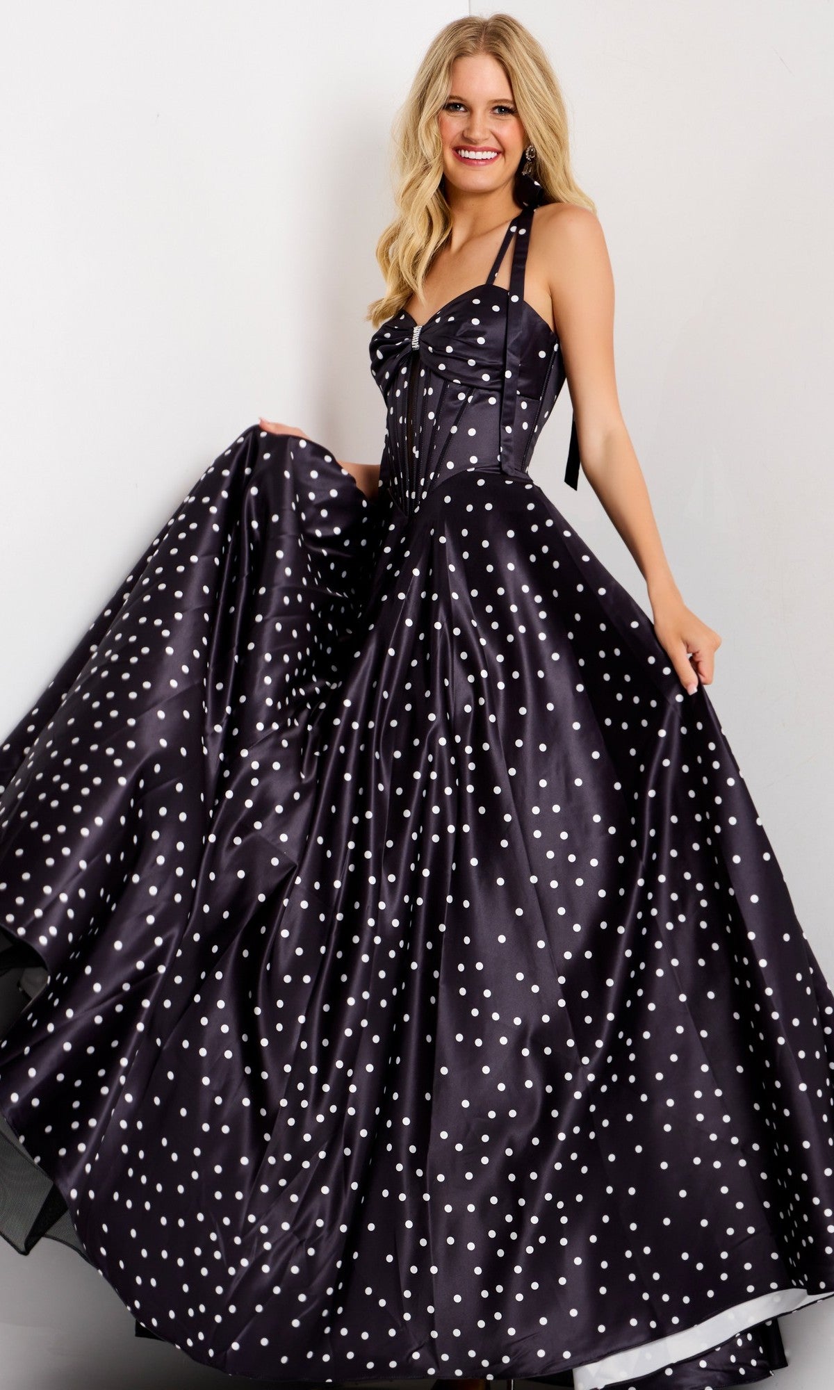 A woman smiles in the Jovani 49774 Long Prom Dress in black, featuring a polka-dot satin fabric, halter neckline, and flowing A-line skirt as she holds out the sides against a plain background.